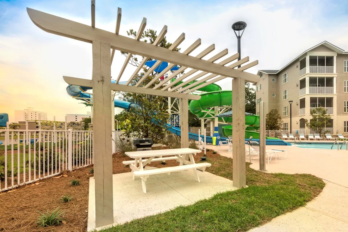 This joyful exterior scene highlights an outdoor area featuring a pergola with a picnic table, a spacious swimming pool, and fun water slides. The space is filled with potential for relaxation, shared meals, and poolside enjoyment, echoing the retreat's playful and romantic promise. Sunlight and recreational elements evoke days spent unwinding together just steps from the beach.