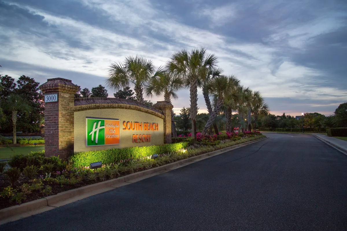 The image showcases the resort’s illuminated sign at its landscaped entrance, bordered by tall palm trees and inviting pathways. The evening lighting and lush greenery create a welcoming first impression, enticing guests to arrive and begin their stay.
