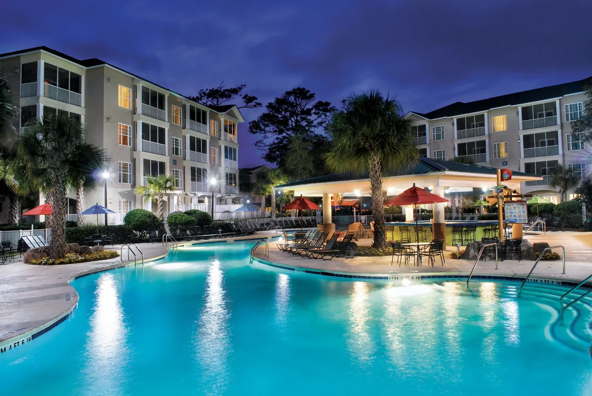 This photo features the resort’s sparkling swimming pool lined with lounge chairs, surrounded by palm trees and an outdoor bar. Guests are seen socializing and relaxing poolside as the sun sets, highlighting the lively community environment.