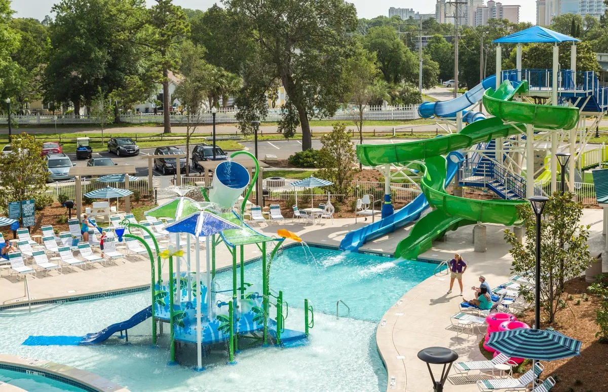 This vibrant exterior image captures the heart of Splash Cove, showcasing twisting water slides, a splash pad, a lively pool, and children joyfully playing in the sun. Lounge chairs line the poolside, emphasizing relaxation, while the energy of family fun is palpable throughout the scene.