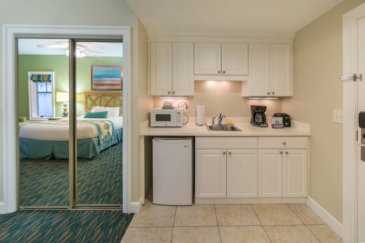 A wide view of the studio space featuring a Queen bed, kitchenette, and a cozy sitting area. Decorative art adds a personal touch while natural light highlights the comfort of both the bed and kitchenette. The arrangement suggests both relaxation and functionality, with the TV visible from the bed and a glimpse of outdoor scenery through a window.