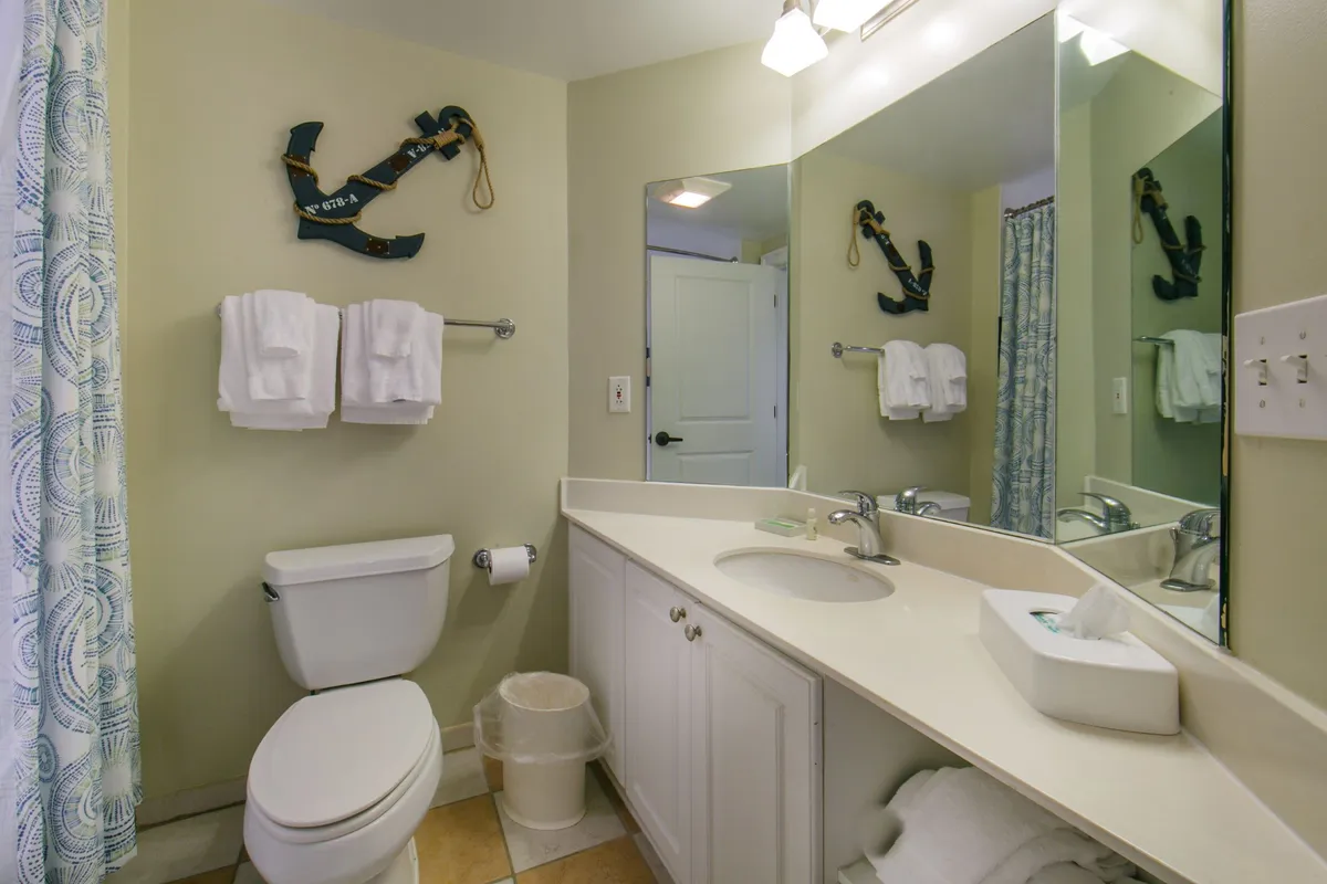 A serene bathroom scene with a rack of fluffy white towels, nautical anchor décor, and a well-lit sink and mirror area. The space feels clean, tranquil, and thoughtfully designed—perfect for morning routines or unwinding after a day at the beach.